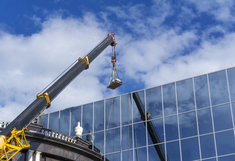 telescopic boom of a construction crane lifts the load against the mirror wall of the building reflecting the sky
