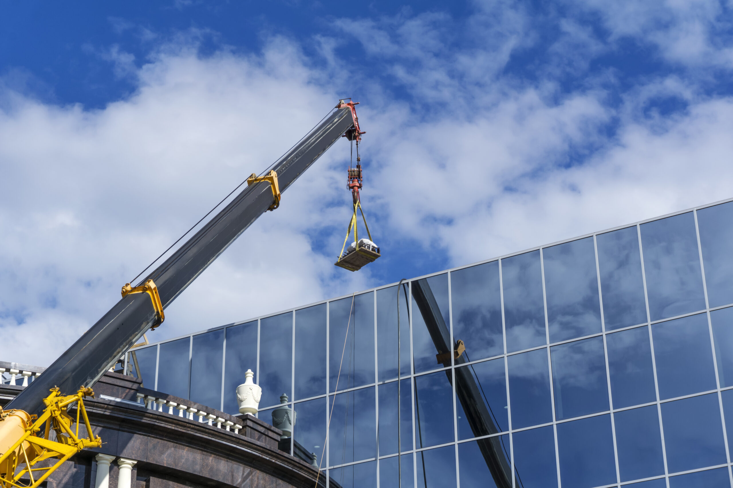 telescopic boom of a construction crane lifts the load against the mirror wall of the building reflecting the sky